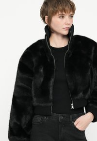 Chaqueta bomber de piel sintética negra con un alto collar y puños acanalados, con cierre de cremallera y bolsillos laterales. Lleva puesta una camiseta negra.