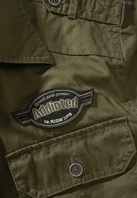 Gros plan sur un tissu vert olive avec une poche boutonnée et un patch noir portant l'inscription "Addicted speed and power to ride 1975".