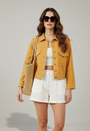 Femme portant de grandes lunettes de soleil noires, veste moutarde à boutons par-dessus un crop top blanc, short blanc, portant un sac tissé à l'épaule.
