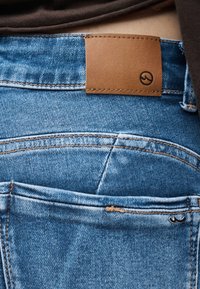 Κοντινή λήψη της πίσω τσέπης μπλε τζιν με καφέ δερμάτινο ένθετο και μικρή μεταλλική λεπτομέρεια λογότυπου στην άκρη της τσέπης.