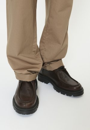 Braune Leder-Schnürschuhe mit dicken schwarzen Sohlen, getragen zu beige umgeschlagenen Hosen vor weißem Hintergrund.