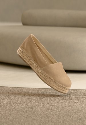 Beige suède espadrille instapschoen met gevlochten jute plateauzool die boven een gestructureerd beige tapijt zweeft.