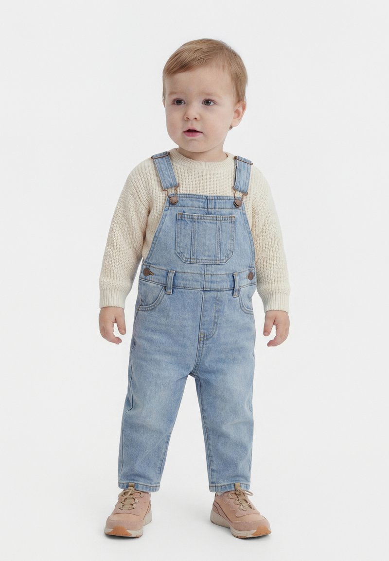 Mazs bērns stāv, ģērbies krēmkrāsas adītā džemperī, gaiši zilās džinsa kombinezonā un rozā sporta apavos uz vienkārša balta fona.