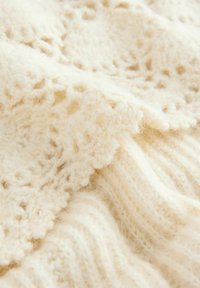 Cremefarvet strikket stof med en blød, tekstureret overflade. Har en kombination af ribbede og blonde mønstre, hvilket øger den visuelle interesse.