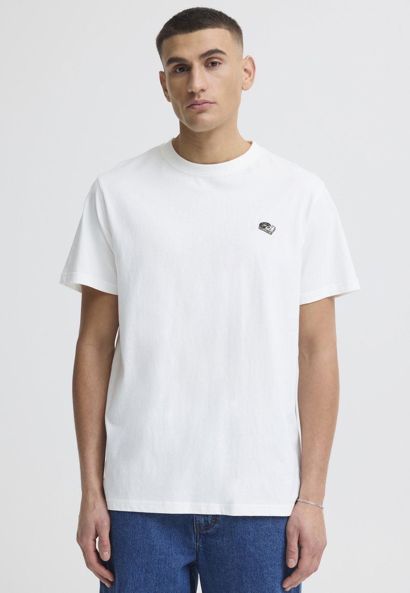 Jeune homme aux cheveux courts et foncés, portant un t-shirt blanc uni avec un petit motif brodé et un jean bleu sur un fond clair.