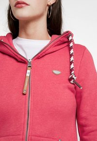 Roze ritshoodie van zachte stof, met een gevlochten trekkoord, metalen ritssluiting en een klein logo-accent op de borst.