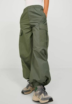 Olivgrüne Cargo-Hosen mit relaxed Fit, ausgestattet mit Seitentaschen, elastischem Bund und leichtem Glanz. Kombiniert mit beigen Sportschuhen.