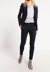 Námořnický modrý blejzr přes světle šedou košili, spárovaný s tmavými kalhotami a šedými kotníkovými botami. Blejzr má upravený střih a jedno knoflíkové zapínání.