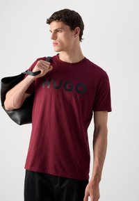 Fiatal férfi bordó HUGO pólót és fekete nadrágot visel, a vállán egy fekete bőr táskával, oldalra néz.