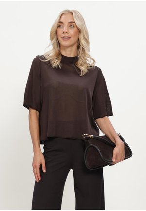 Blonde Frau trägt ein locker sitzendes schwarzes Kurzarmtop und schwarze Hose, hält eine schwarze Handtasche mit goldenen Details und schaut zur Seite.