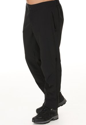 Schwarze Softshell-Hosen mit glatter Textur, ausgestattet mit seitlichen Reißverschlusstaschen und einem taillierten Bein Design, kombiniert mit schwarzen Wanderstiefeln.