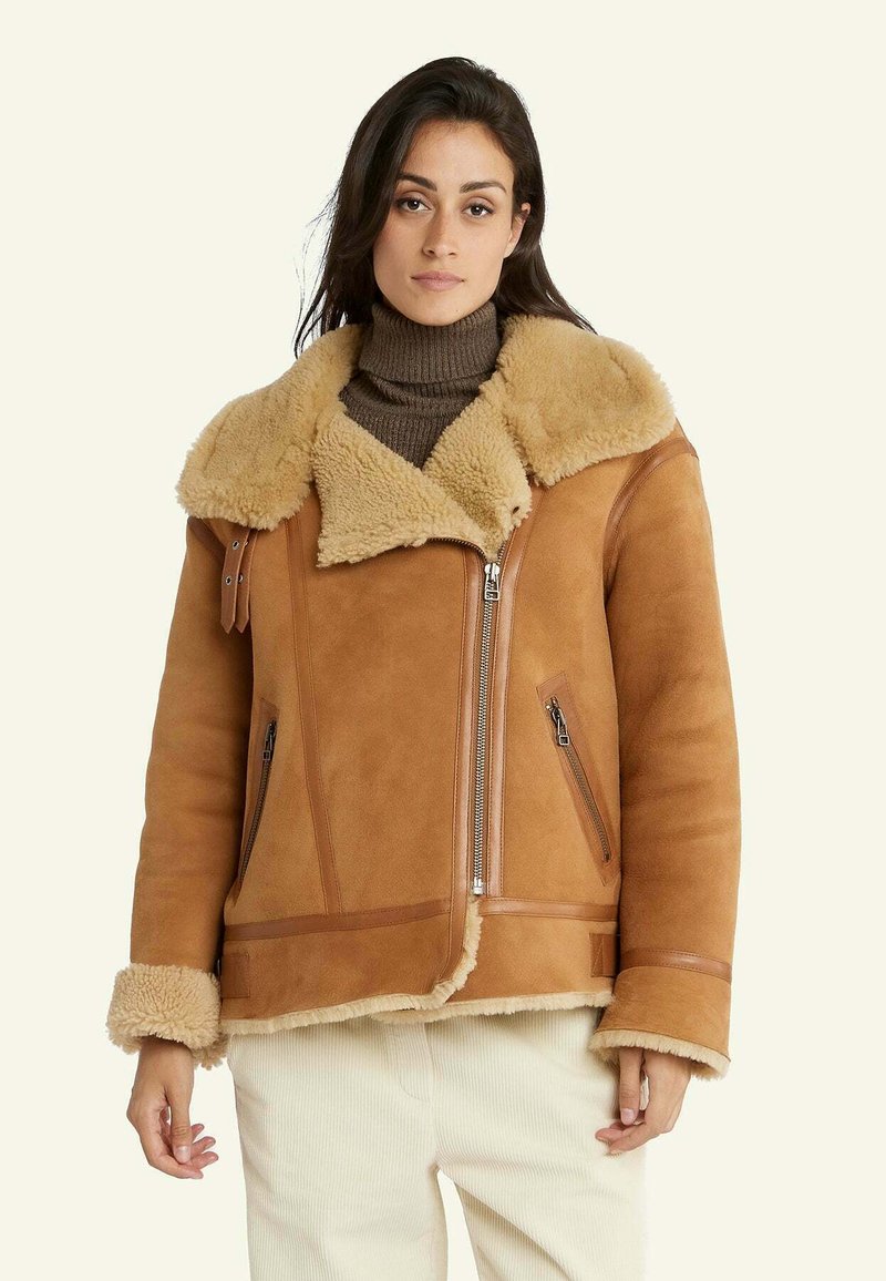 Tansheepskin jack met een grote kraag, ritssluiting aan de voorkant en twee zijzakken met ritssluiting. Heeft contrasterende bruine leren accenten. Zachte textuur.