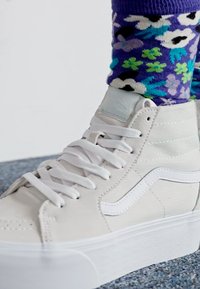 Witte hoge sneakers met een gladde leren bovenkant, witte veters en een gestructureerde zool. Kleurrijke sokken met bloemenpatroon zichtbaar boven de schoen.