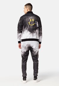 Chándal temático del espacio en negro y gris con patrones de nubes y estrellas, que presenta una cara sonriente amarilla y un gráfico de astronauta en la parte posterior de la chaqueta.