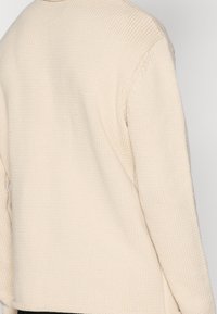Pull en maille beige avec une finition texturée, doté d'un col rond et de manches tombantes. Le design est décontracté avec des fentes sur les côtés en bas.