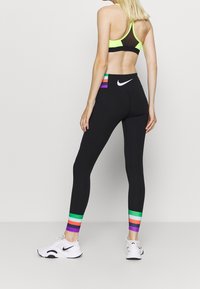 Czarne legginsy z wielokolorowymi paskami na kostkach, w zestawieniu z czarnym stanikem sportowym w neonowym zielonym kolorze. Widoczne logo Nike na legginsach.