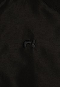 Črno blago z subtilno teksturo, z majhnim črnim izvezenim logotipom na spodnjem levem kotu. Preprost, minimalističen dizajn.