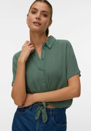 Femme portant une chemise verte à manches courtes boutonnée et nouée à la taille, un jean bleu, les cheveux tirés en arrière et des boucles d'oreilles argentées en forme d'anneau.