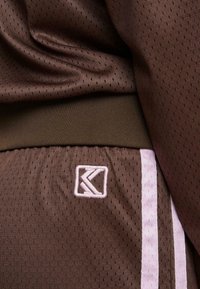 Rudas sportinis audinys su tinkluotu raštu. Yra siuvinėta rožinė juosta ir šviesiai rožinė logotipo etikete šone.