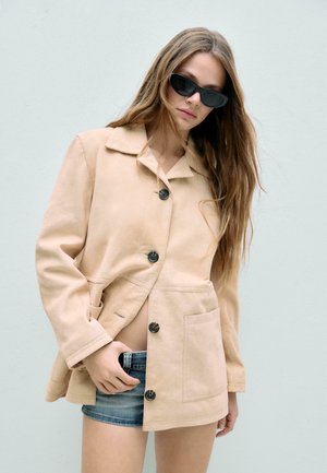 Kvinde med langt hår iført sorte solbriller, beige jakke med knapper og denimshorts, stående mod en ensfarvet lys baggrund.