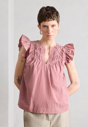 Person med kort hår iført en lyserød tekstureret top med flæseærmer og beige bukser, stående mod en ensfarvet lys baggrund.