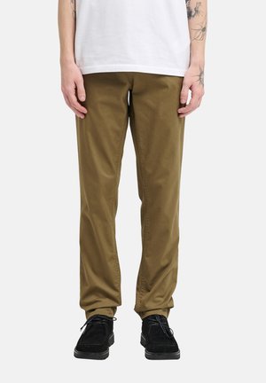 Homme portant un pantalon vert olive, des chaussures noires à lacets, une chemise blanche ; tatouages visibles sur les poignets des deux bras sur un fond uni.