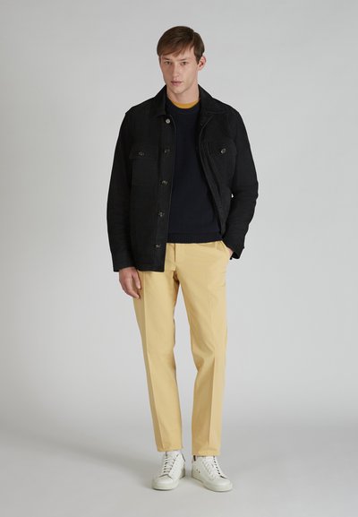 Czarna kurtka z płótna z dwoma kieszeniami na piersi, noszona na granatowym swetrze. Beżowe spodnie i białe trampki dopełniają stylizację.
