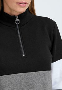 Černá mikina na zip s vysokým límcem, která má šedé a bílé horizontální barevné bloky, je vyrobena z bavlny a má stříbrný detail zipu.