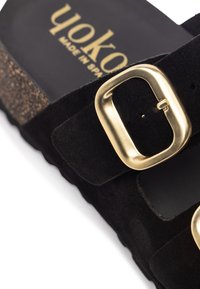 Infradito in suede nero con suola in sughero, dotate di una grande fibbia in metallo dorato e del nome del marchio "Yoko" impresso sulla soletta.