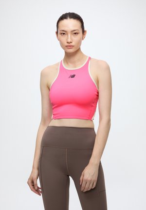 Femme aux cheveux foncés attachés, portant un crop top rose New Balance et des leggings marron taille haute, debout sur fond blanc.