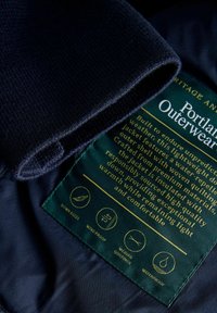 Jūras spēku ārējā jaka ar mīkstu, teksturētu apkakli un auduma etiķeti, kurā ir norādītas kopšanas instrukcijas un laikapstākļiem izturīgas ikonas.