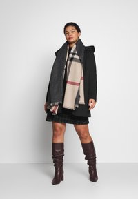 Melnas kapuces mētelis, bēša un pelēka kvadrātu šalle, kvadrātu svārki un burgundijas krāsas augstās zābaki līdz ceļiem. Strukturēts dizains ar teksturētiem audumiem.