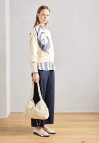 Sudadera beige con gráfico de un pájaro azul, superpuesta a una camisa de rayas. Pantalones de pierna ancha oscuros y zapatos crema con hebilla. Sosteniendo un bolso tote claro.