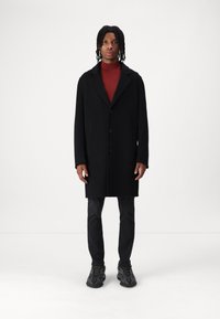 Sort uldfrakke med notch-krave, frontknapper og sidelommer, lagdelt over en rød turtleneck, parret med mørke jeans og sneakers.
