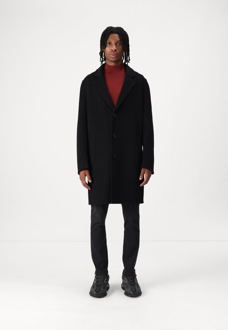 Sort uldfrakke med notch-krave, frontknapper og sidelommer, lagdelt over en rød turtleneck, parret med mørke jeans og sneakers.