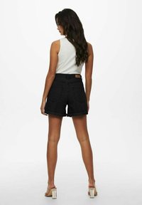 Pantalones cortos de mezclilla negra con dobladillos enrollados, con bolsillos traseros y una etiqueta marrón en la cinturilla, combinados con una blusa sin mangas blanca.