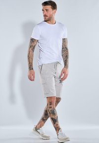 White crew-neck t-shirt, light gray cargo shorts with pockets, and beige/white sneakers. Intricate arm tattoos are visible.