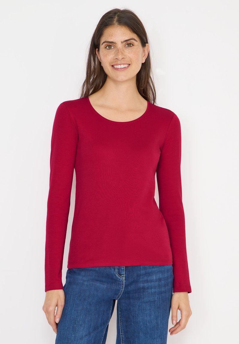 Maglia rossa a maniche lunghe in un tessuto liscio ed elasticizzato con scollatura a barchetta, abbinata a jeans blu. Design semplice, senza motivi o decorazioni.