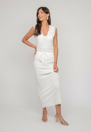 Femme aux longs cheveux foncés souriante, portant une robe blanche sans manches avec une taille à cordon et des talons dorés à lanières, debout devant un fond uni.