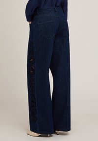 Jeans de mezclilla de pierna ancha en un lavado oscuro, con un intrincado diseño de flores recortadas en negro a lo largo del costado, bolsillos traseros estándar y costuras cosidas.