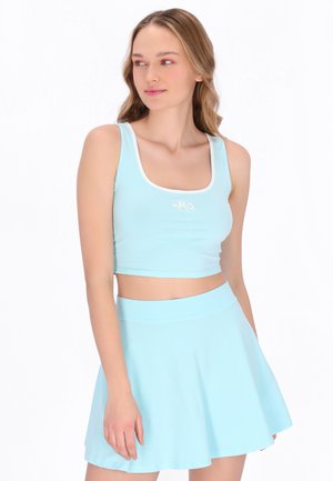 Hullámos hajú fiatal nő világos kék, haspóló és hozzáillő magasderekú szoknya viseletében, oldalra nézve.