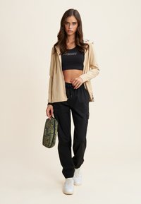 Sudadera con cremallera color beige claro, top corto negro con el logo "FRDDY", pantalones cargo negros, zapatillas blancas y un bolso verde con estampado.