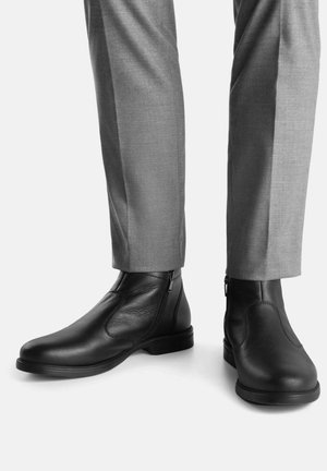 Črni usnjeni gležnjarji z bočnimi zadrgami, gladka tekstura, okrogel nos, kombinirani s sivimi, krojenimi hlačami. Osredotočenost na elegantno obliko in prileganje.