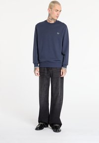 Sweatshirt blu navy con polsini e scollatura a costine, abbinato a jeans in denim scuro a gamba larga e mocassini neri. Presente un semplice logo.