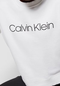 Vit sweatshirt med ribbad halsringning, med texten "Calvin Klein" i svart font över framsidan. Mjuk tyg, avslappnad design.