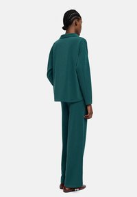 Zestaw loungewear w kolorze teal, składający się z luźno dopasowanej bluzki z długim rękawem i kołnierzykiem oraz spodni z szerokimi nogawkami. Gładka tekstura, minimalistyczny design, brak widocznych wzorów.