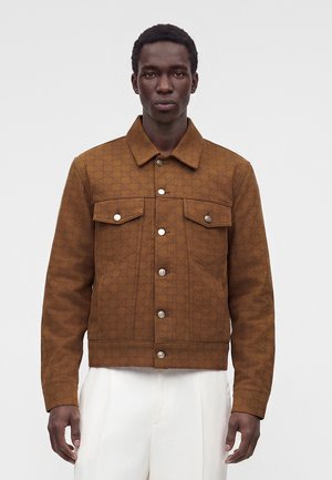 Homme portant une veste marron boutonnée avec deux poches poitrine et un pantalon blanc, debout devant un fond clair uni.