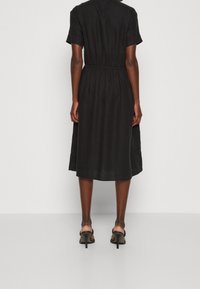 Černé šaty délky po kolena s krátkými rukávy, elastickým pasem a hladkou texturou. Nošeny s černými sandály na podpatku. Jednoduchý a minimalistický design.