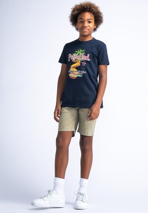Marine T-shirt met kleurrijke ananasafbeelding en "Petrol Ind." tekst, gecombineerd met beige shorts met kleine palmbomenpatronen. Witte sneakers.