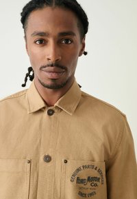 Hnědá košile na knoflíky vyrobená z odolné látky, s dvěma předními kapsami a potiskem loga na levé kapse. Klasický design límce.